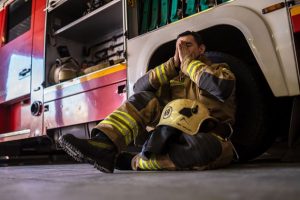Firefighter sitting against fire truck with head in hands