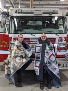 Firefighters Mike Skinner and Daniel Sundahl hold up quilts in front of fire truck