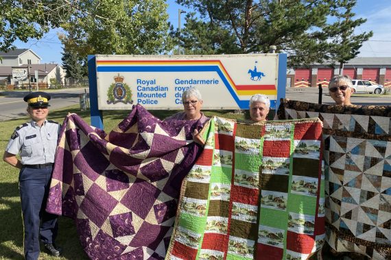 RCMP members stand with Patti-Jo Healey holding up quilts in front of RCMP sign